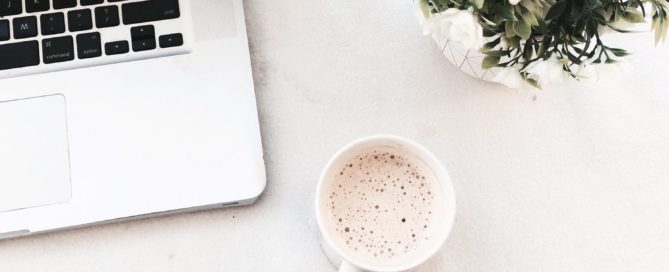 person holding a hot drink next to a laptop and a vase of flowers
