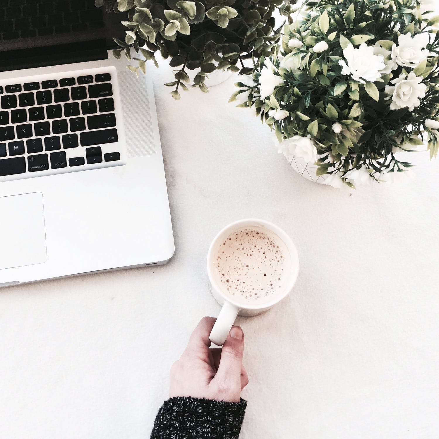 person holding a hot drink next to a laptop and a vase of flowers